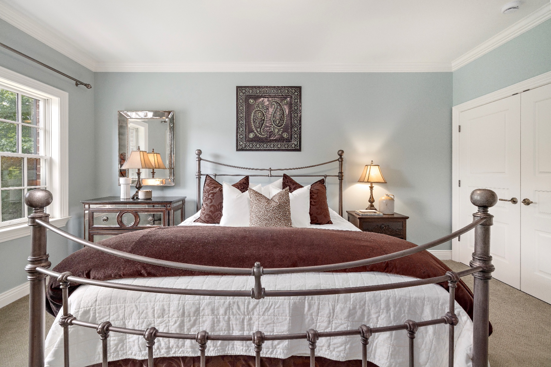 Guest bedroom with iron four-poster bed, soft blue walls, mirrored nightstand, and window to the trees
