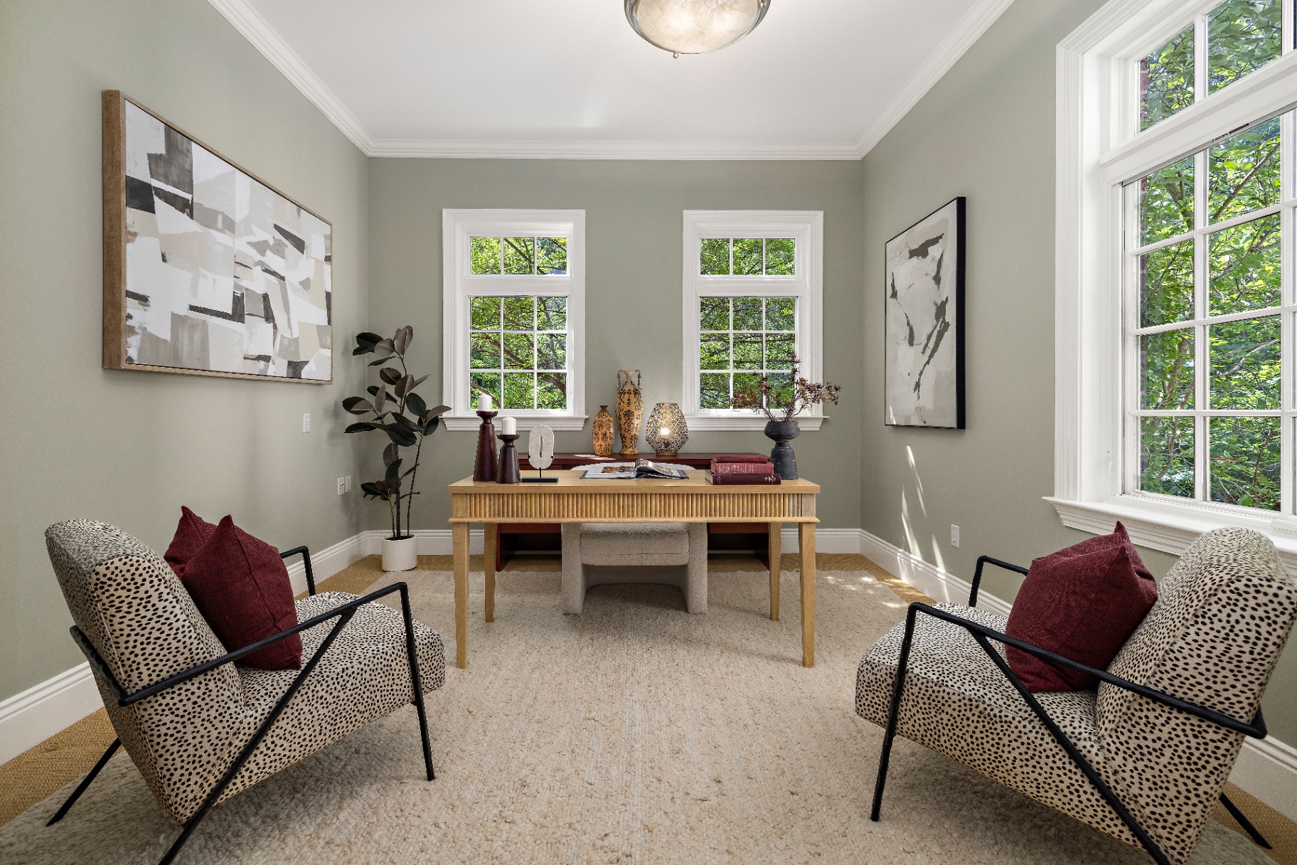 Main floor office with sage green walls, writing desk, patterned accent chairs, and tall windows to the trees